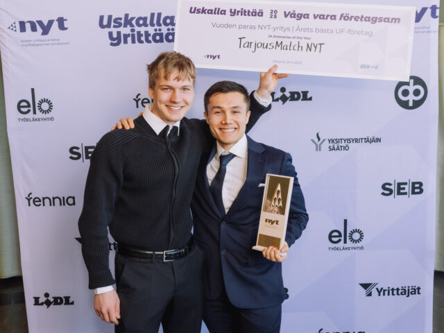 Kaksi henkilöä hymyilee kameralle kuvausseinän edessä, toisella kädessä pokaali ja toinen pitelee plakaatia päiden yläpuolella.